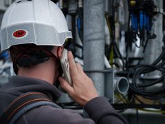 Worker in a hard hat talks on a phone beside industrial pipes and cables, showing maintenance, safety, communication, and technical work in a factory setting