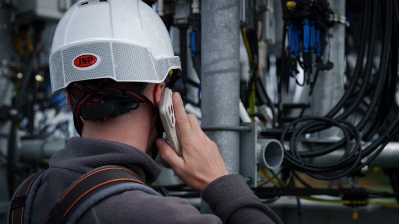 Worker in a hard hat talks on a phone beside industrial pipes and cables, showing maintenance, safety, communication, and technical work in a factory setting