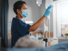 Hospital Ward: Professional Black Head Nurse Wearing Face Mask Does Checkup of Patient's Vitals, Checking Heart Rate Computer, Intravenous or Iv Fluids Drip Bag. Caring Nurse Monitors Person Recovery